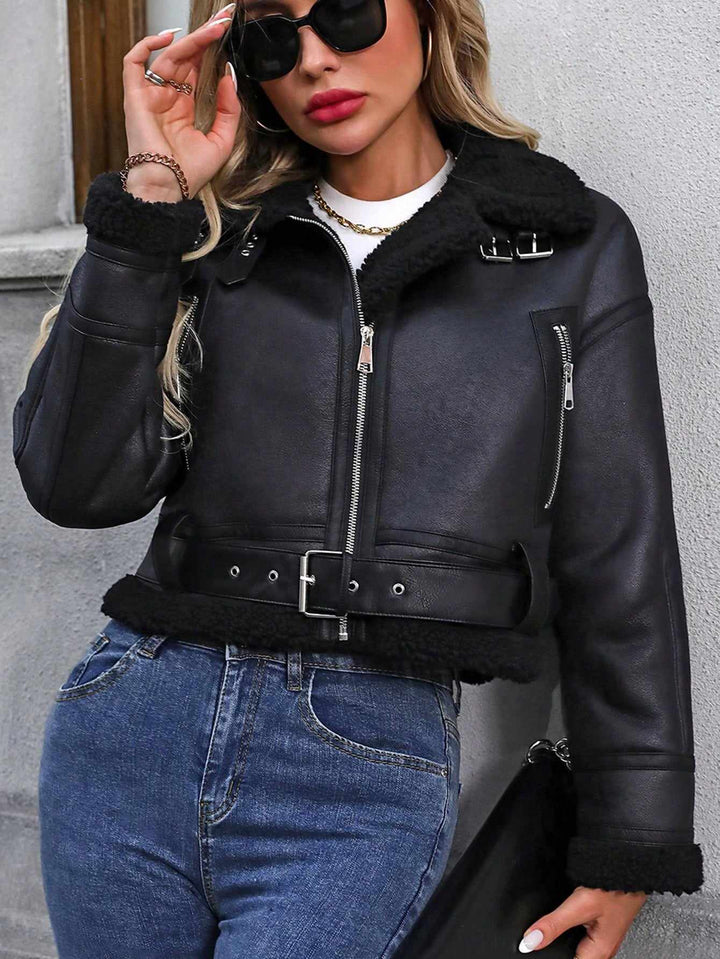 Woman wearing a black leather jacket with fur trim and blue jeans against a gray wall.