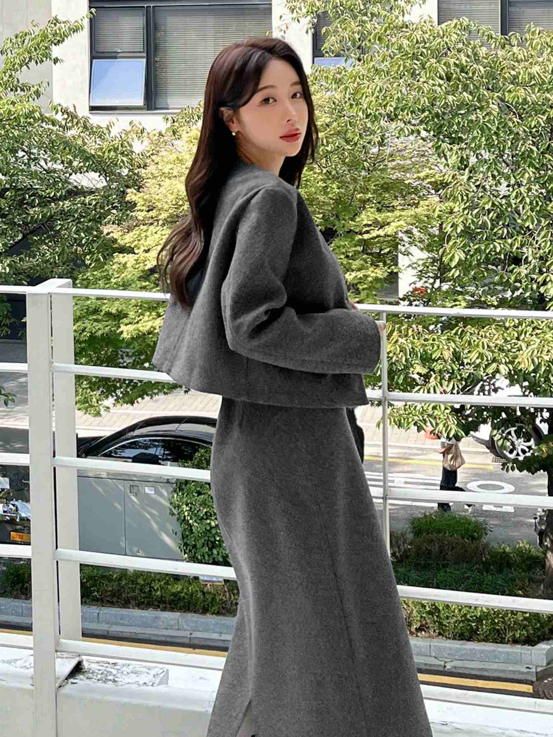 A woman wearing a dark gray cropped jacket and matching long skirt, standing on an outdoor balcony with trees and a street in the background.