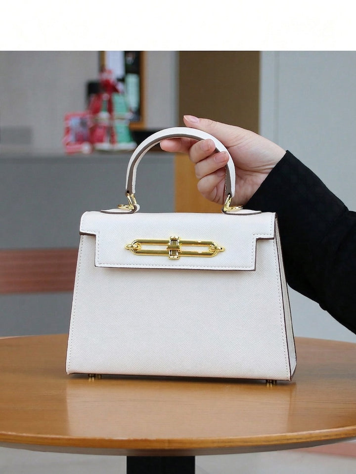 A hand holding a structured white leather handbag with a gold turn-lock clasp and top handle, set on a wooden table.