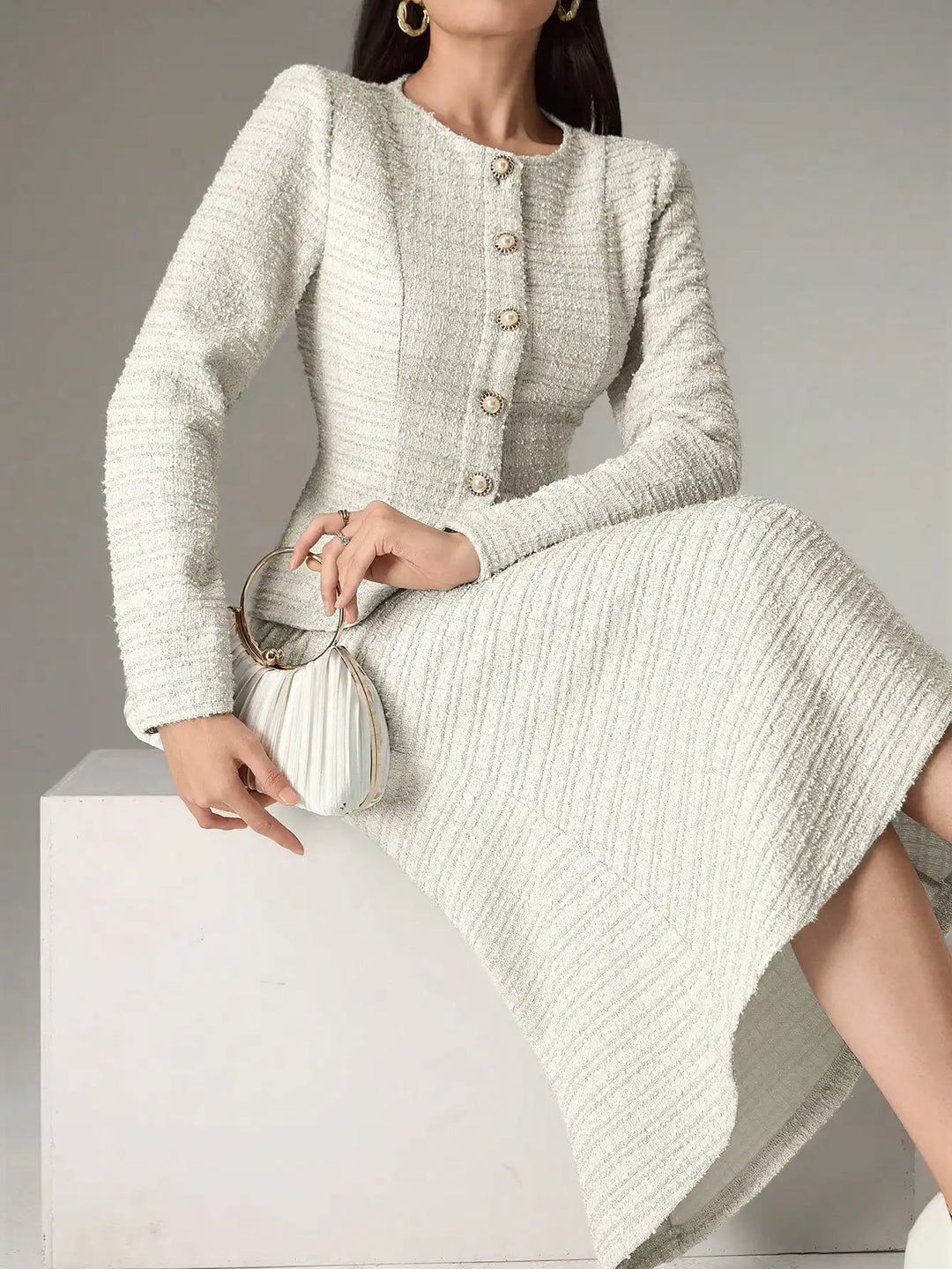 A woman models a light beige textured tweed long-sleeve dress with decorative buttons down the front, paired with a matching pleated clutch bag.