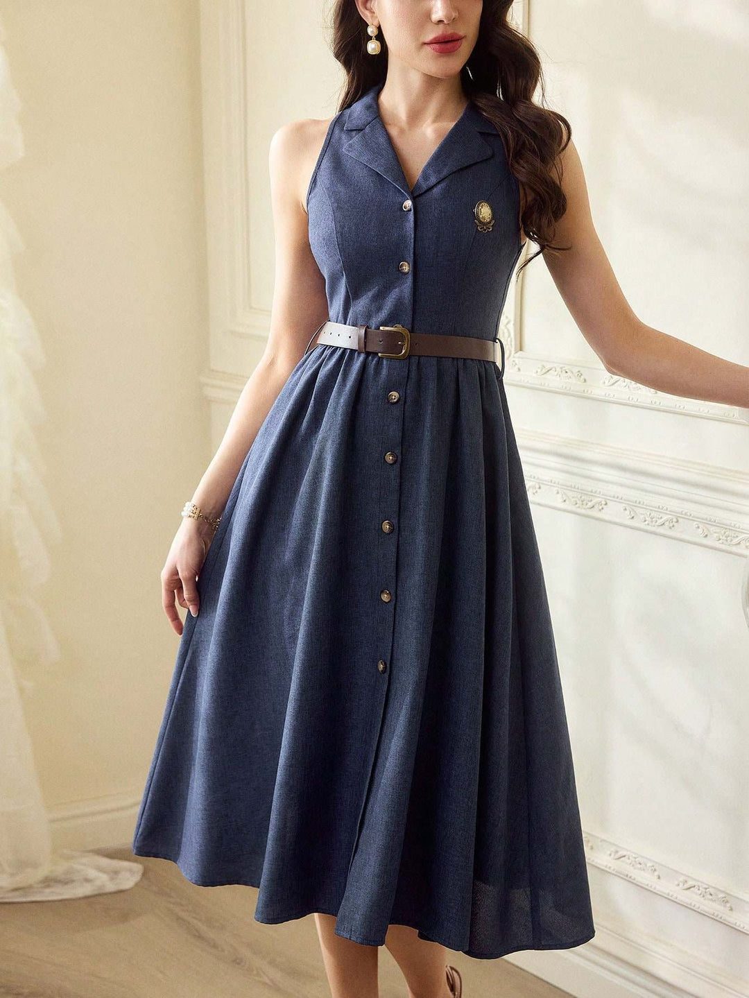A woman wearing a sleeveless navy blue midi dress with a button-front bodice, a brown belt, and a small gold emblem on the chest, standing in an indoor room with cream-colored walls.