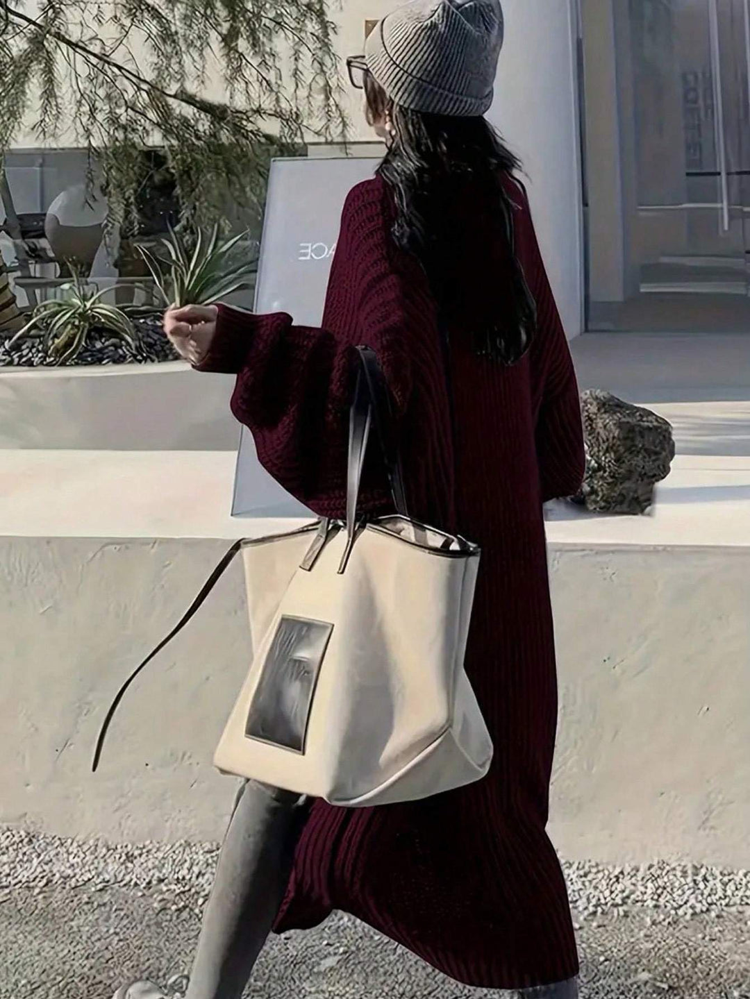 A person wearing a ribbed burgundy knit cardigan, gray beanie, and glasses carries a large cream canvas tote bag with black handles and a black front patch, walking outdoors near a modern building.