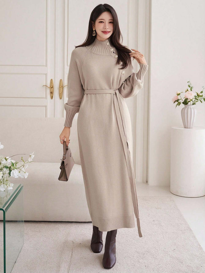 A woman models a beige knit midi dress with a mock neck, long sleeves, and a matching tie belt. She is standing in a bright, modern room with a white door, a white sofa, and decorative plants.