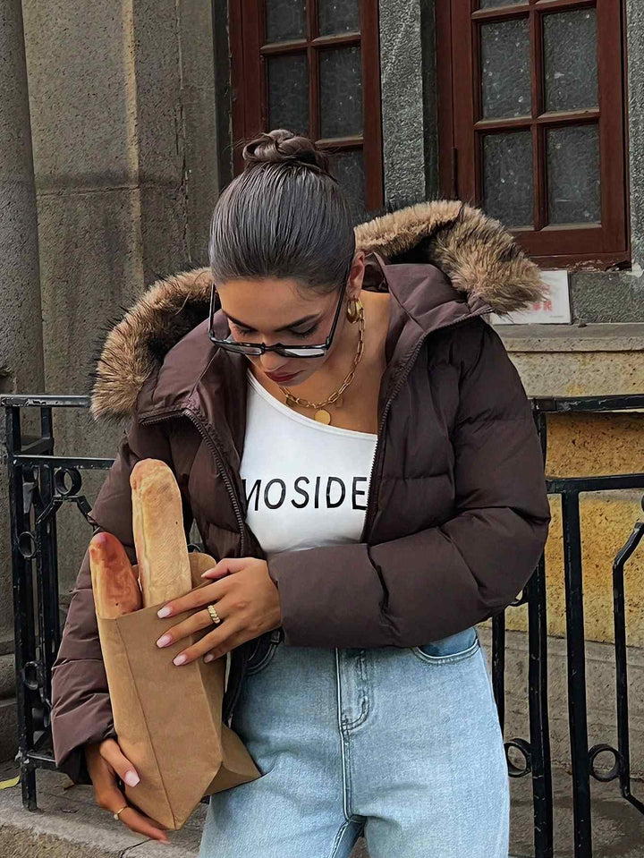A woman wearing a dark brown puffer jacket with a faux-fur trimmed hood, a white tank top with &