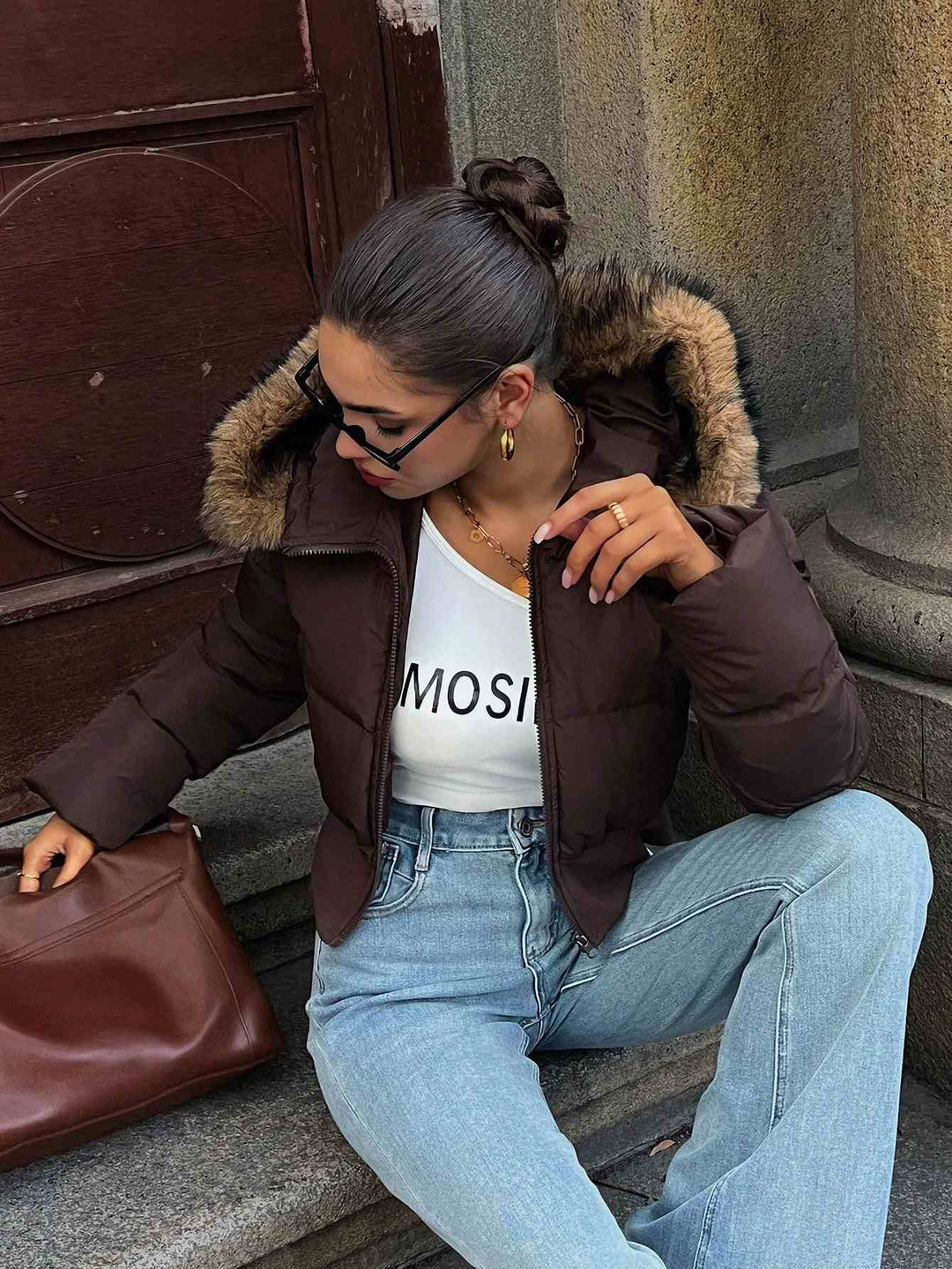 A woman sits on stone steps wearing a dark brown puffer jacket with a faux-fur trimmed hood over a white top and light blue jeans, holding a brown leather handbag.