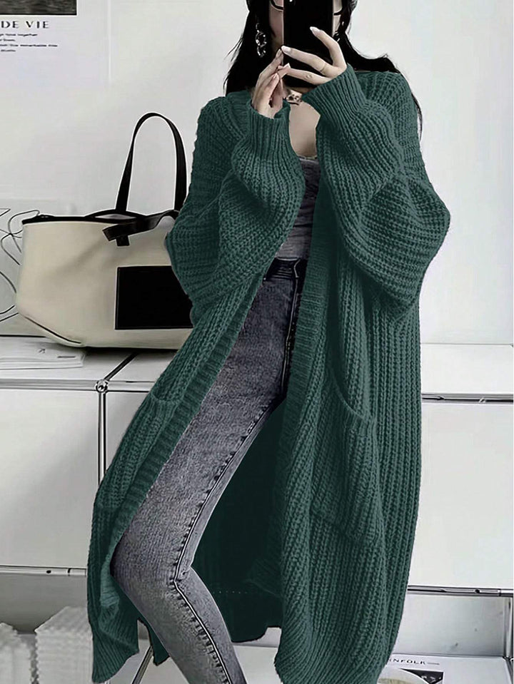 A woman wearing an oversized, long-sleeved dark green chunky knit cardigan over a gray top and gray jeans, standing in a room with a white bag on a shelf in the background.