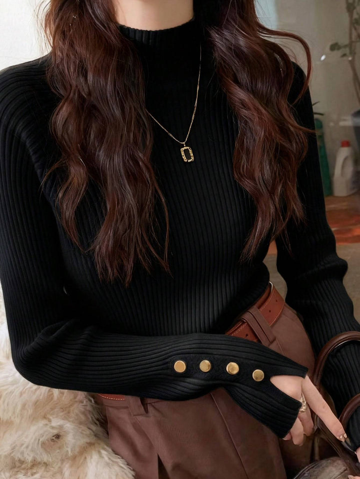 A woman wearing a black ribbed mock-neck long-sleeve sweater with gold button details on the cuffs, paired with brown pants and a gold pendant necklace.