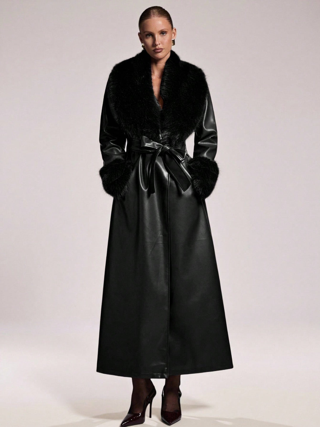 A model wearing a long black faux-leather coat with black fur trim and a matching black belt, paired with dark pointed-toe heels, against a light gray studio background.