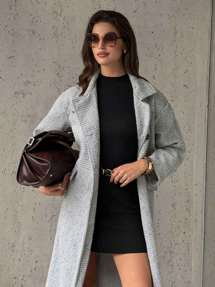 A woman models a light gray textured overcoat over a black dress, holding a dark brown leather handbag against a textured gray concrete wall.