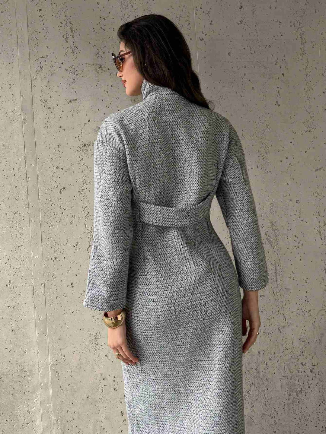 A woman shown from the back wearing a textured light gray wrap coat with a matching belt, standing against a concrete wall.