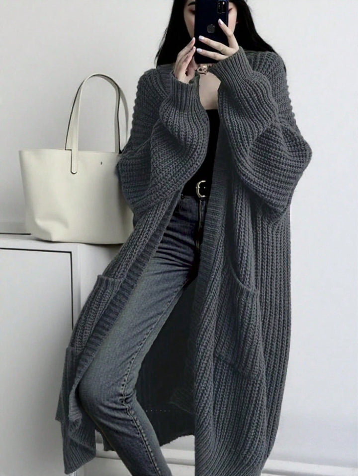 A woman takes a mirror selfie wearing a long, chunky ribbed gray cardigan over a black top and gray jeans. A white tote bag sits on a white cabinet to her left against a white wall.