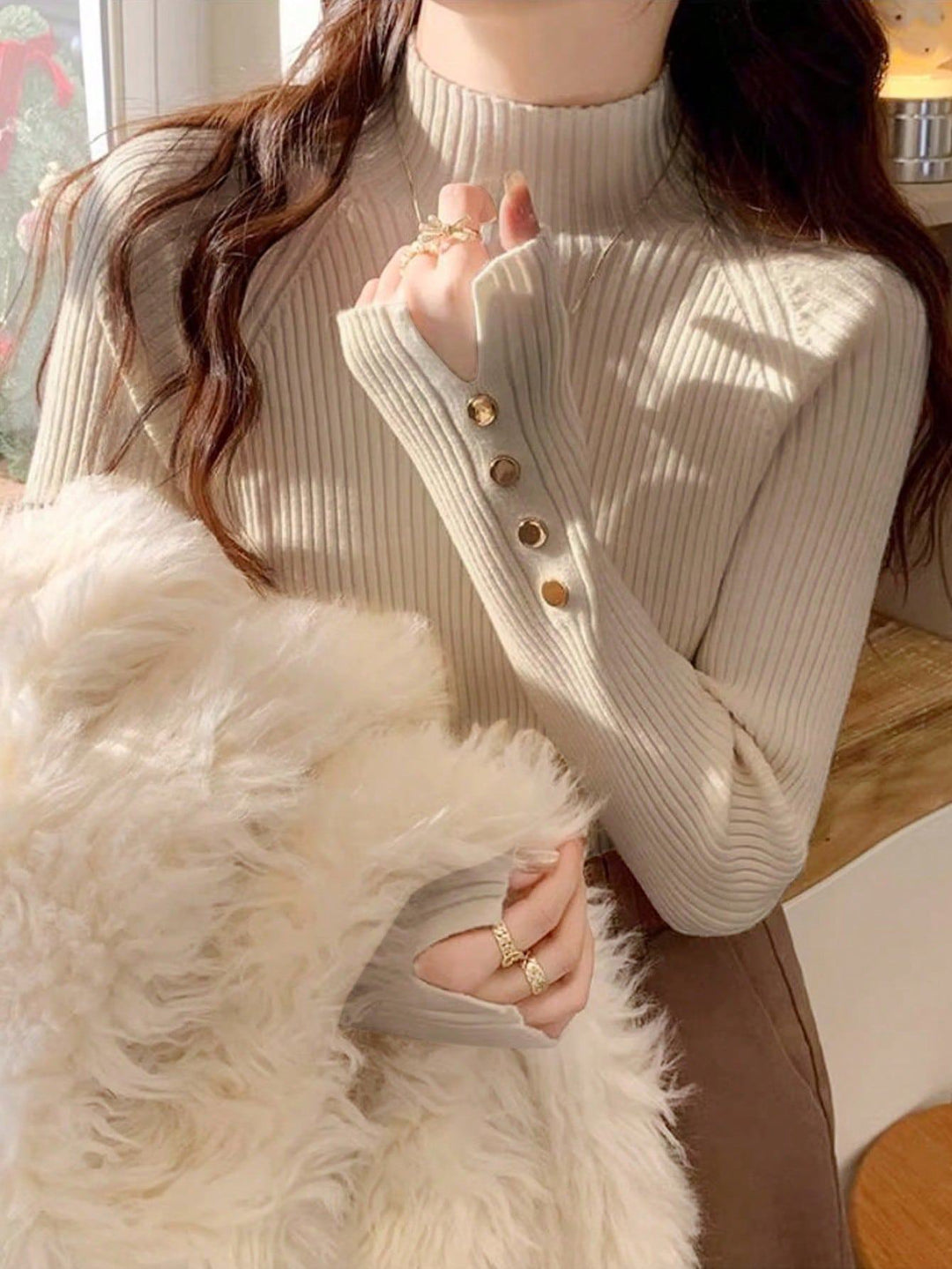 Person wearing a beige ribbed sweater with button details, holding a fluffy white pillow.