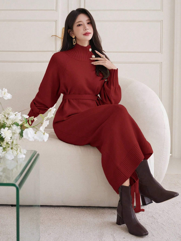 A woman sits on a cream-colored curved sofa wearing a long-sleeve, high-neck, ribbed knit dress in a deep burgundy red. The dress features a matching tie belt at the waist and a tie detail at the hem.