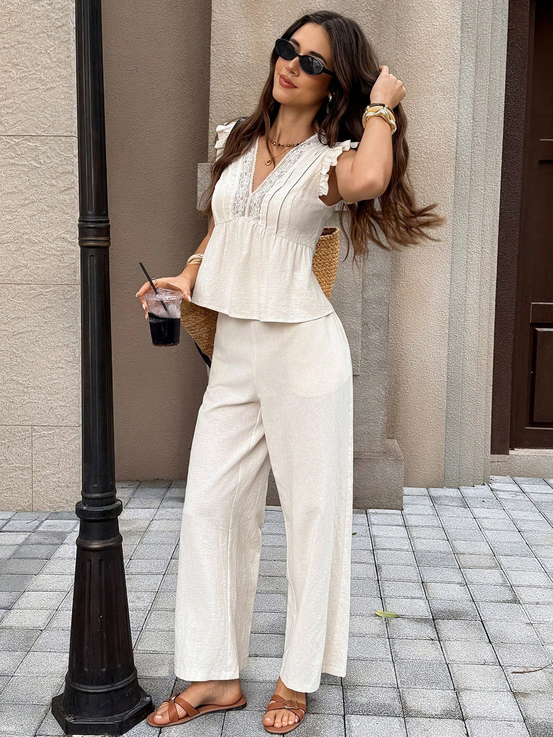 A woman models a two-piece cream linen set consisting of a ruffled short-sleeve top and matching wide-leg trousers, styled with sunglasses, a straw bag, and flat sandals in an outdoor urban setting.