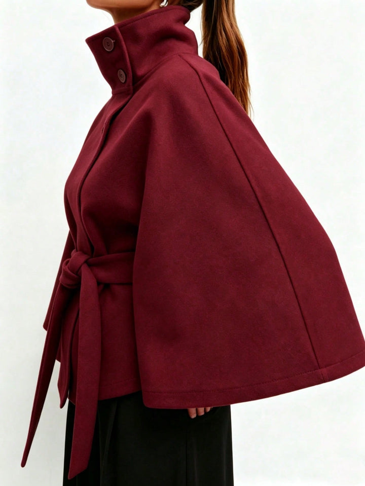 A close-up side view of a model wearing a burgundy cape-style coat with a high collar, buttoned neckline, and self-tie belt, paired with black bottoms against a plain white background.