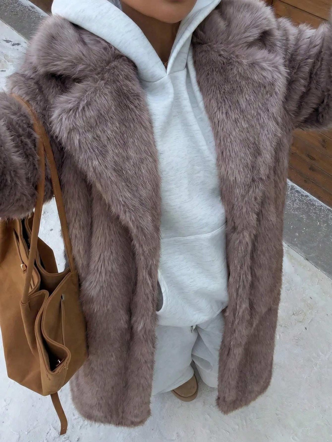 A person wearing a long taupe faux fur coat over a white hoodie and sweatpants, carrying a tan shoulder bag in an outdoor snowy setting.