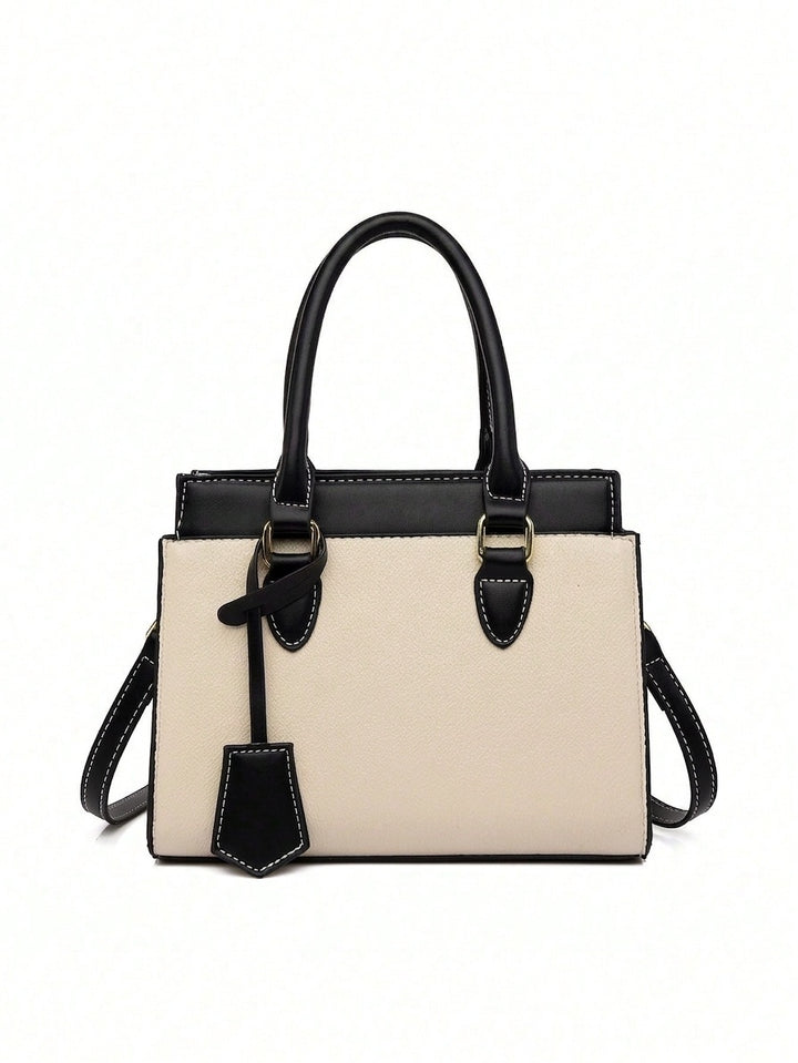A structured tote handbag with a cream-colored front body and black leather handles, trim, and strap, featuring gold-tone hardware and a hanging leather tag, set against a plain white background.