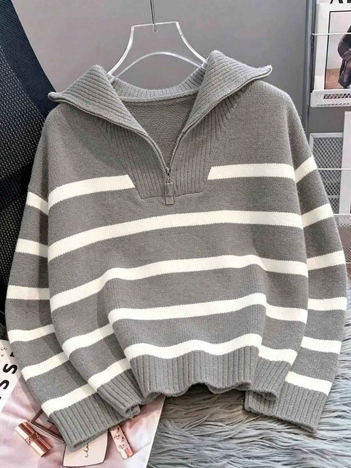 A gray and white horizontally striped knit sweater with a mock neck and short front zipper, hanging on a clear hanger against a gray background.