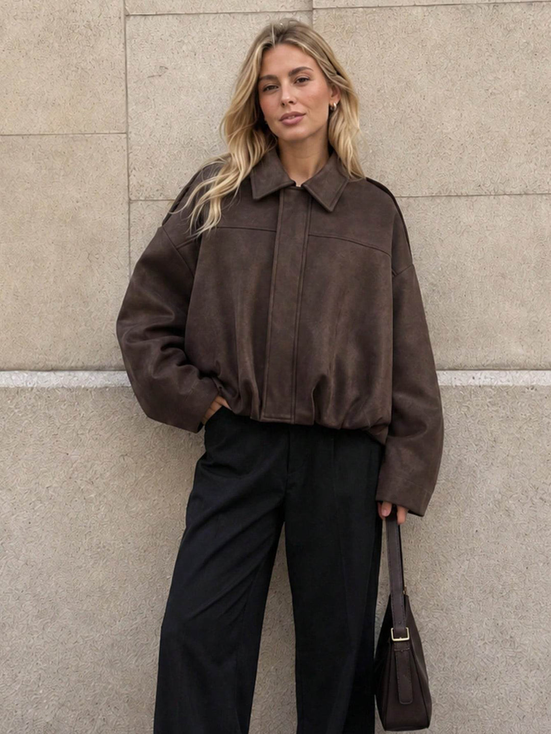 A woman stands against a light beige stone wall wearing a dark brown cropped suede jacket, black wide-leg trousers, and holding a dark brown shoulder bag.