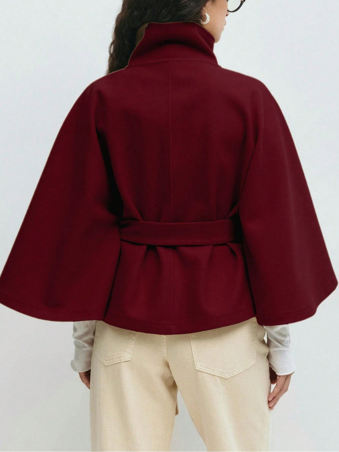 A person shown from the back wearing a deep burgundy cape-style wrap jacket with a high collar and self-tie waist belt, paired with light beige pants.