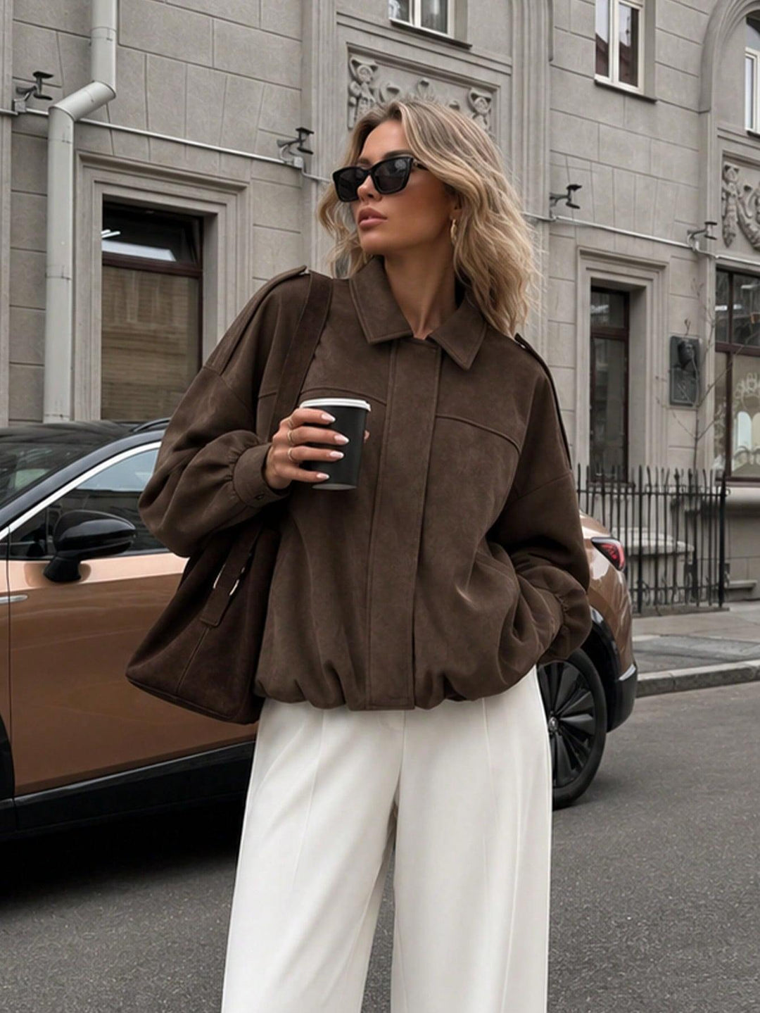 A woman wearing a brown oversized suede jacket and white wide-leg pants stands on a city street holding a black coffee cup. She is wearing black sunglasses and carrying a brown shoulder bag.
