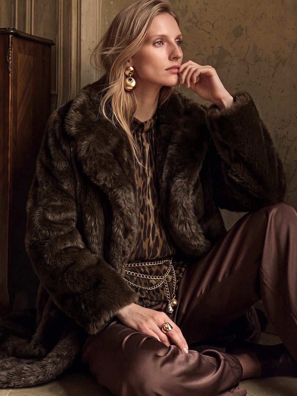 A woman sits indoors wearing a dark brown fur coat over a leopard-print top and brown satin pants, with layered gold jewelry and a chain belt. The setting has a textured beige wall and a dark wooden cabinet.