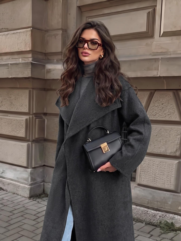 A woman wearing a long charcoal gray wool coat, black turtleneck, black handbag with gold hardware, and glasses stands in front of a stone building.