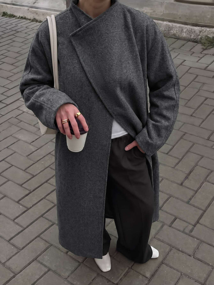 A person wearing a long, dark gray wrap-style coat over a white top and dark wide-leg pants, standing on a gray brick pavement. They are holding a white cup and wearing a beige shoulder bag.